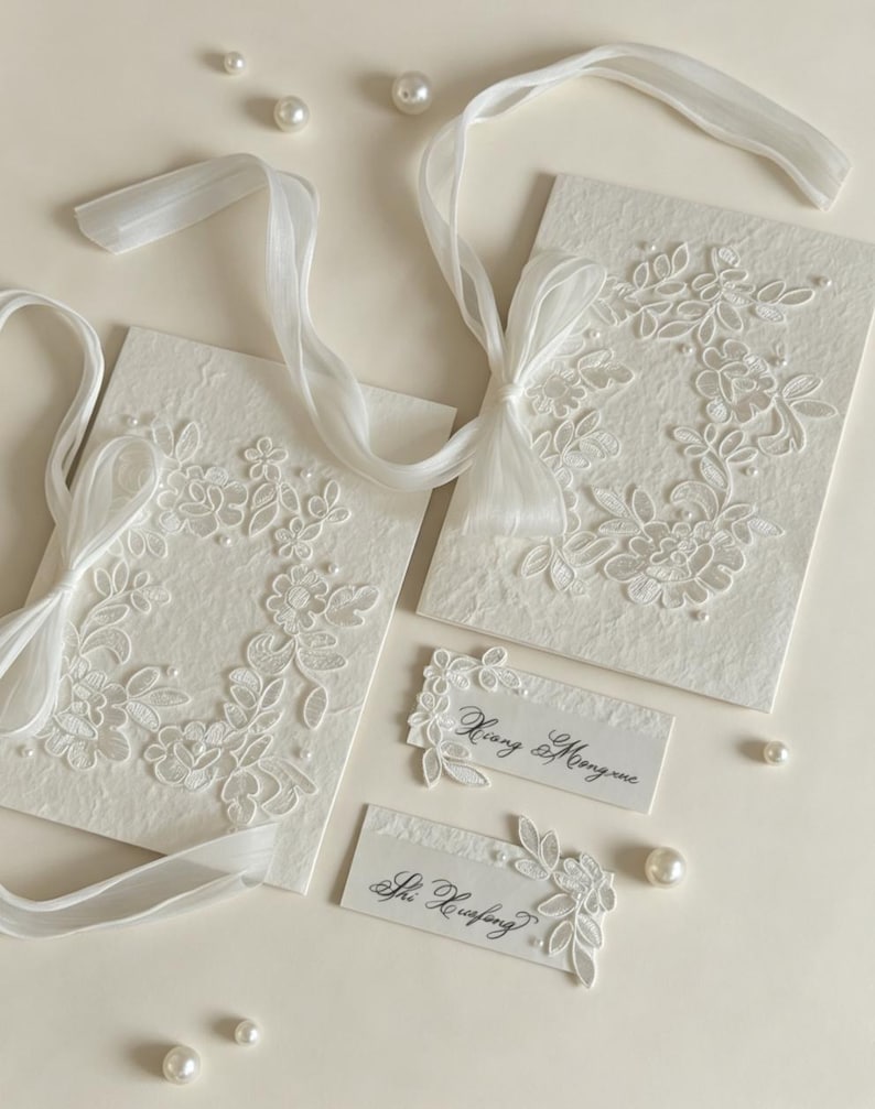 May include: Two off-white textured paper cards featuring white floral lace appliqu&eacute;s, sheer white ribbon bows, and small pearl accents. Two smaller rectangular cards are included, one with the text Xiang & Mengxue and one with Shi Xuefeng.