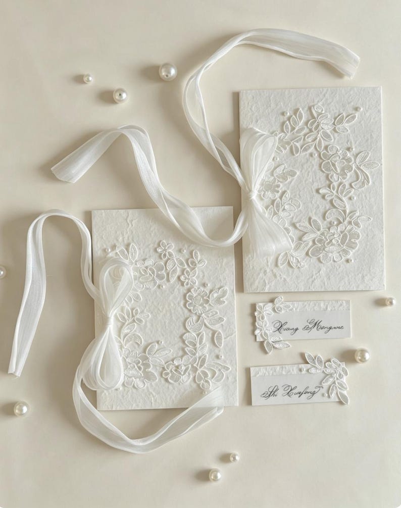 May include: Two rectangular white textured paper cards featuring white floral lace appliqués, small pearl accents, and long sheer white ribbons tied in bows. Two smaller rectangular name cards with black cursive text and lace details are included.