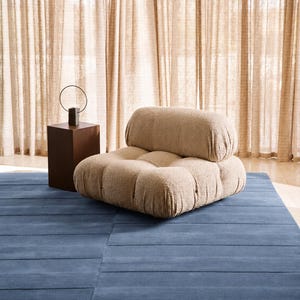 May include: A low-profile, beige, textured boucl&eacute; armchair with a tufted seat and backrest, positioned next to a dark brown rectangular side table that holds a thin, circular metal sculpture on a small, dark, rectangular base.