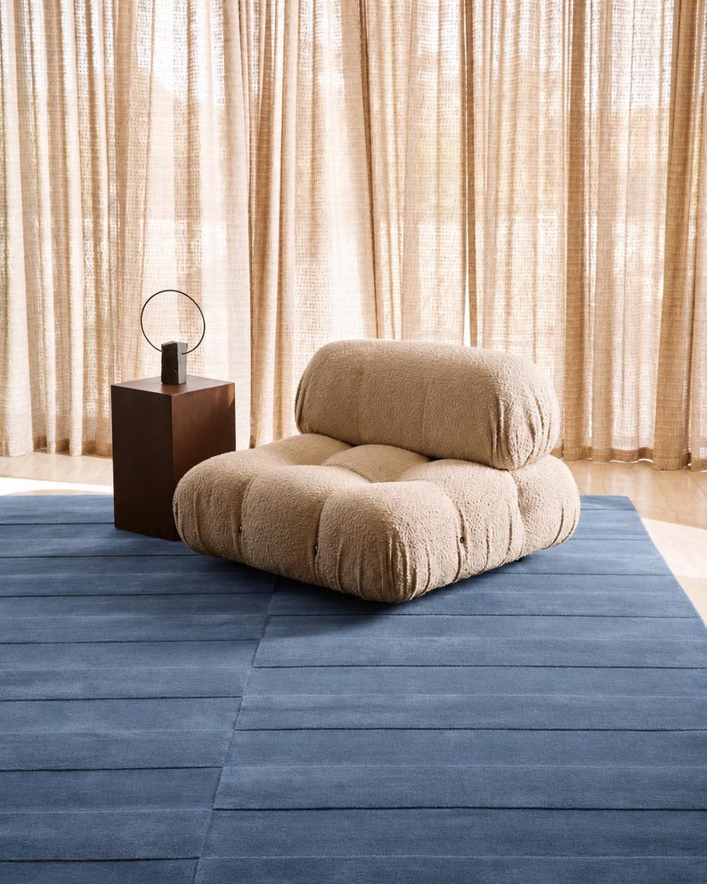 May include: A low-profile, beige, textured boucl&eacute; armchair with a tufted seat and backrest, positioned next to a dark brown rectangular side table that holds a thin, circular metal sculpture on a small, dark, rectangular base.