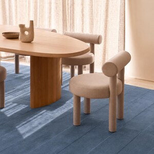 May include: A light wood oval dining table with a central pedestal base, accompanied by several upholstered chairs in a soft beige fabric. The chairs feature a cylindrical backrest and four tubular legs. A small ceramic vase sits on the table.