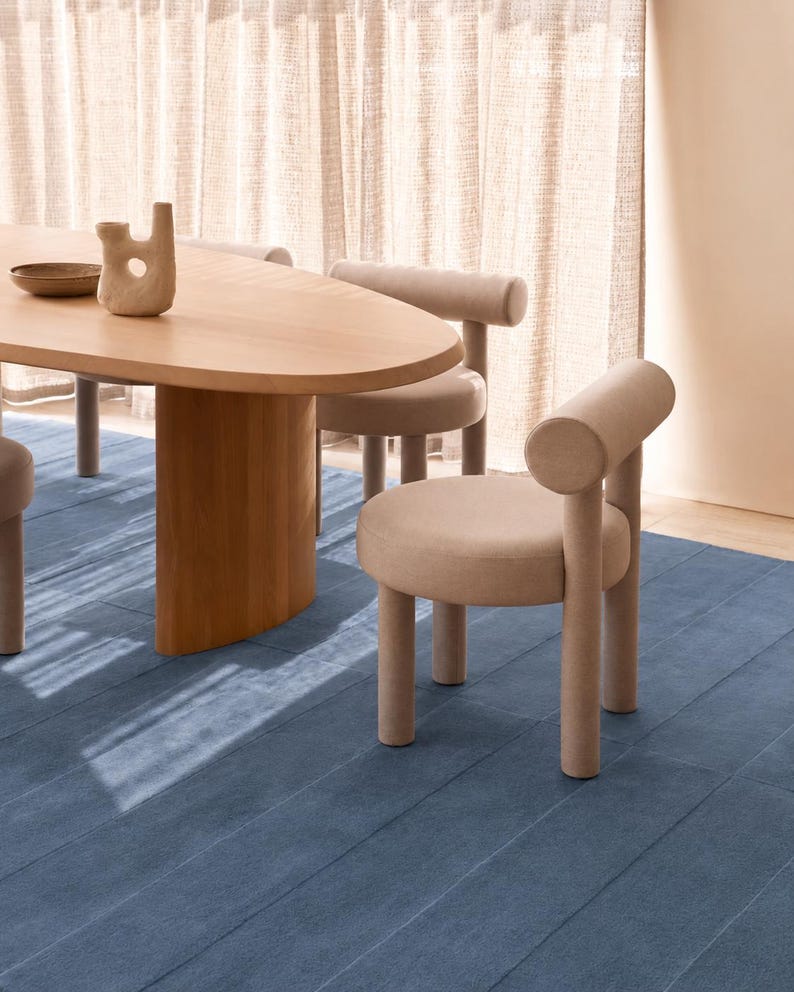 May include: A light wood oval dining table with a central pedestal base, accompanied by several upholstered chairs in a soft beige fabric. The chairs feature a cylindrical backrest and four tubular legs. A small ceramic vase sits on the table.