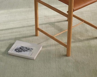 May include: A light green rug with a wooden chair and a book. The chair has a brown leather seat and back. The book has a black and white image on the cover. The chair is made of light-colored wood.