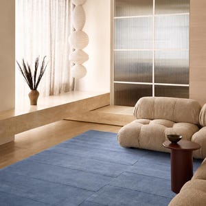 May include: A rectangular blue area rug with a linear grid pattern, featuring a textured surface. The rug is positioned on a light-colored floor in a room with a beige modular sofa, a dark brown cylindrical side table, and a vertical paper lantern.