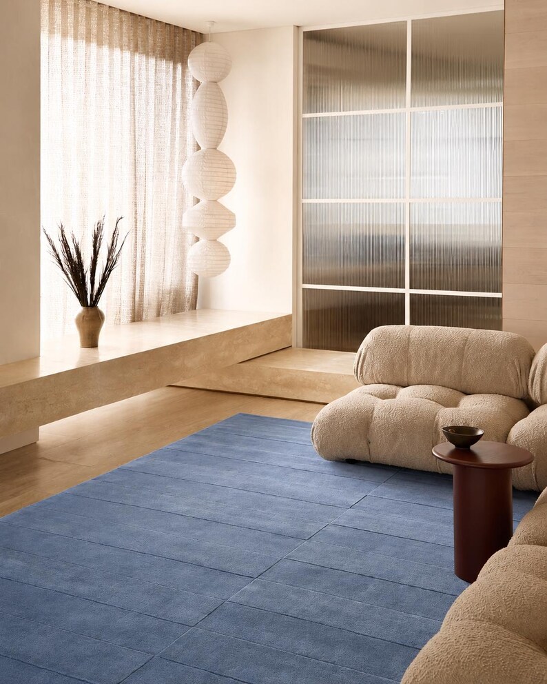 May include: A rectangular blue area rug with a linear grid pattern, featuring a textured surface. The rug is positioned on a light-colored floor in a room with a beige modular sofa, a dark brown cylindrical side table, and a vertical paper lantern.