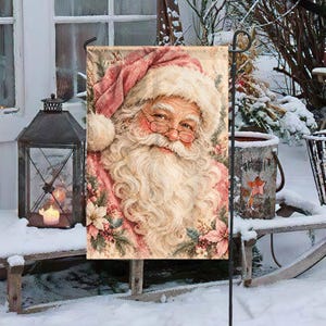 May include: A rectangular garden flag featuring a detailed illustration of Santa Claus with a long white beard, wearing a red hat and glasses. The design includes accents of pink poinsettia flowers and greenery, set against a soft, muted background.