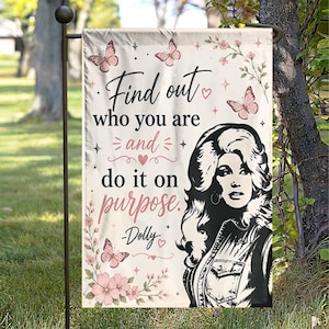 May include: A rectangular garden flag featuring a black and white illustration of a woman with voluminous hair, pink butterflies, and floral accents. The text reads: Find out who you are and do it on purpose. -Dolly. The background is off-white.