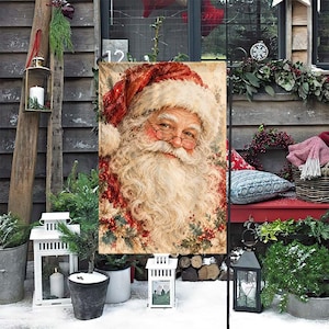 May include: A rectangular garden flag featuring a detailed illustration of Santa Claus with a white beard, wearing a red hat and glasses. The design includes holly berries and green leaves at the bottom, set against a textured, vintage-style background.