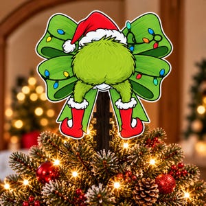 Peut inclure: Une décoration de sapin de Noël représentant un personnage vert et poilu portant un bonnet de Père Noël rouge et des bottes rouges, placé devant un grand nœud vert orné d'une guirlande lumineuse colorée, fixé sur un piquet vertical noir.