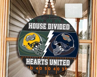 May include: A round wooden door sign featuring a football field background with the text HOUSE DIVIDED at the top and HEARTS UNITED at the bottom. It displays a yellow Green Bay Packers helmet and a navy Chicago Bears helmet separated by a white lightning bolt.
