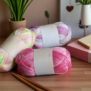 Op de afbeelding: Drie bollen kleurrijk garen in roze, paarse en groene tinten, elk omwikkeld met een blanco etiket. Twee houten breinaalden liggen op de voorgrond. Het garen is gerangschikt op een houten oppervlak.