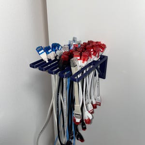 May include: A navy blue 3D-printed cable organizer mounted on a white wall, holding multiple network cables. The cables have red, white, blue, and black connectors. The organizer is designed to keep cables neat and accessible.