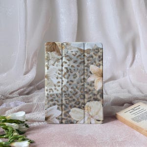 Puede incluir: Funda para tableta con diseño floral y de leopardo en tonos neutros. La funda tiene una cubierta plegable y una mezcla de colores beige, marrón y blanco. El diseño incluye flores y un estampado de leopardo.