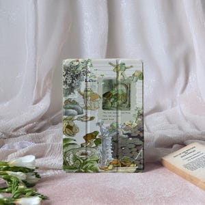 Puede incluir: Una funda para tableta con una ilustración vintage de ranas en un estanque con nenúfares y la palabra "Lilie". El diseño tiene una estética de cuento de hadas con verdes, marrones y cremas.