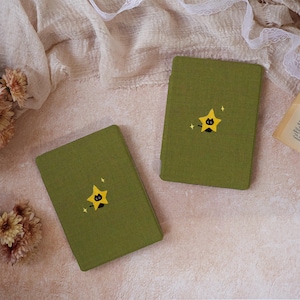 Peut inclure: Deux &eacute;tuis rectangulaires pour liseuse en tissu vert olive textur&eacute;. Chaque &eacute;tui pr&eacute;sente un motif centr&eacute; d'un petit chat noir sortant de derri&egrave;re une &eacute;toile jaune &agrave; cinq branches, agr&eacute;ment&eacute; de deux petites &eacute;toiles blanches &agrave; quatre branches.