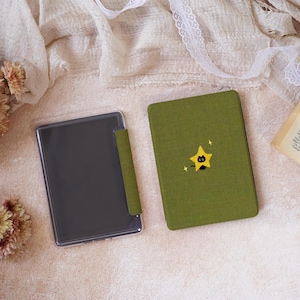 Peut inclure: Un &eacute;tui pour liseuse rectangulaire vert olive avec une finition en tissu textur&eacute;. La couverture avant pr&eacute;sente un petit motif brod&eacute; d'un chat noir assis dans une &eacute;toile jaune avec de minuscules &eacute;tincelles blanches. Une coque de protection transparente est fix&eacute;e.