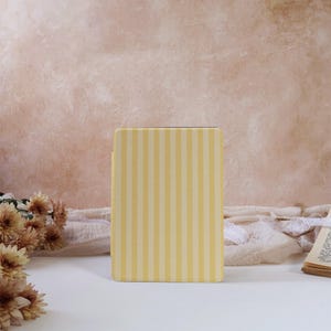 May include: A rectangular book or journal cover featuring a vertical striped pattern in alternating shades of pale yellow and cream. The object is positioned upright, displaying a clean, flat surface with consistent, parallel lines throughout the design.