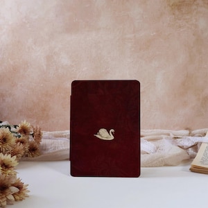 May include: A rectangular, deep burgundy book cover featuring a subtle, embossed floral pattern across its surface. Centered on the front is a small, detailed, cream-colored swan emblem, providing a distinct contrast against the dark red background.
