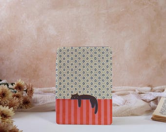 Puede incluir: Un cuaderno rectangular con un diseño estampado. La mitad superior presenta un fondo beige con patrones florales azules. La mitad inferior tiene rayas verticales rojas y rosas, con una ilustración de un gato negro. El cuaderno está sobre una superficie blanca.