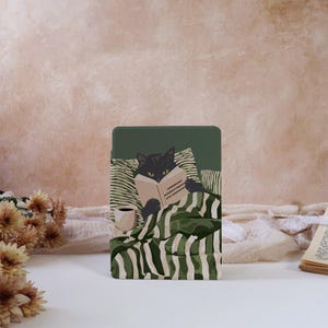 May include: A rectangular e-reader case featuring a dark green background with an illustration of a black cat lying in bed under striped green and white blankets, reading a book titled Comment Domestiquer Les Humains, with a cup of coffee nearby.