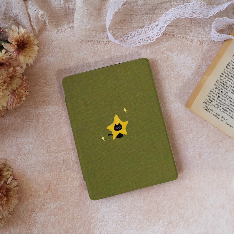 Peut inclure: Une housse de liseuse rectangulaire vert olive orn&eacute;e d'une petite broderie repr&eacute;sentant un visage de chat noir sortant d'une &eacute;toile jaune, agr&eacute;ment&eacute;e de deux minuscules &eacute;tincelles blanches en forme d'&eacute;toile au-dessus et &agrave; gauche.