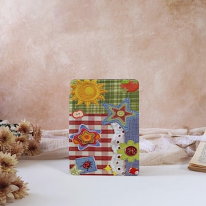 May include: A decorative card featuring a patchwork design with a sun, stars, and a ladybug. The card has a red and white checkered pattern, green plaid, and denim-like fabric. The card is approximately 4 inches wide and 6 inches tall.