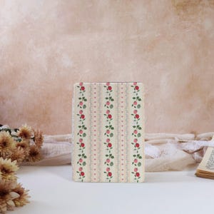 May include: A rectangular, cream-coloured case featuring a repeating pattern of vertical stripes, pink hearts, and floral vines with red and green berries. The case is likely for a tablet or e-reader.