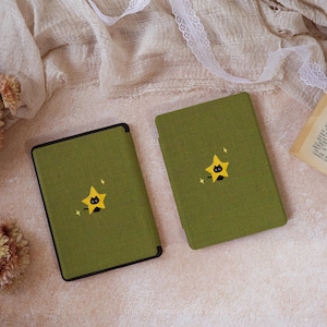 Peut inclure: Deux &eacute;tuis rectangulaires pour liseuse en tissu vert olive textur&eacute;. Chaque &eacute;tui pr&eacute;sente un motif brod&eacute; centr&eacute; repr&eacute;sentant un petit chat noir sortant de derri&egrave;re une &eacute;toile jaune &agrave; cinq branches, agr&eacute;ment&eacute; de deux petites &eacute;toiles blanches.