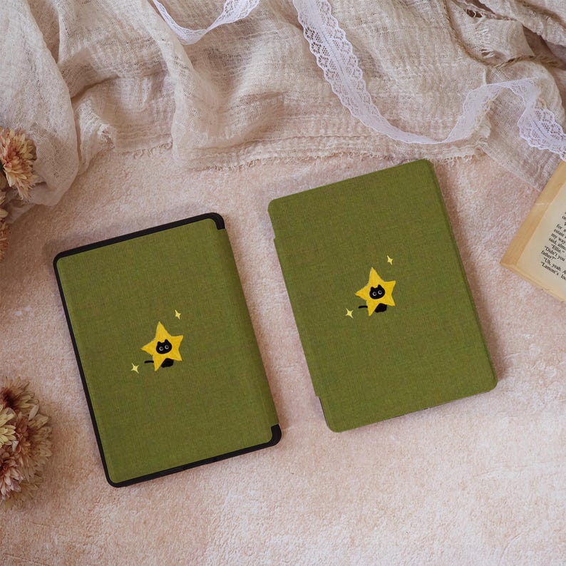 Peut inclure: Deux &eacute;tuis rectangulaires pour liseuse en tissu vert olive textur&eacute;. Chaque &eacute;tui pr&eacute;sente un motif brod&eacute; centr&eacute; repr&eacute;sentant un petit chat noir sortant de derri&egrave;re une &eacute;toile jaune &agrave; cinq branches, agr&eacute;ment&eacute; de deux petites &eacute;toiles blanches.