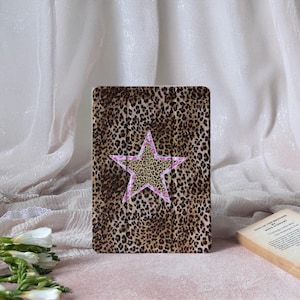 Puede incluir: Una funda rectangular con un diseño de estampado de leopardo y un acento en forma de estrella. La estrella tiene un centro con estampado de leopardo y un contorno rosa. La funda es probablemente para una tableta o un lector electrónico.