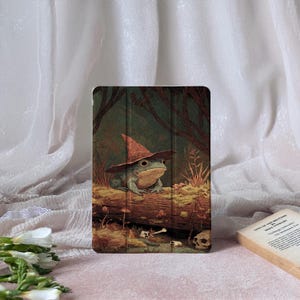 Puede incluir: Funda para tableta con una ilustración fantástica de una rana con sombrero de bruja, ambientada en un bosque. La obra de arte incluye setas, calaveras y un fondo detallado con árboles y follaje. La funda tiene una cubierta verde oscuro.