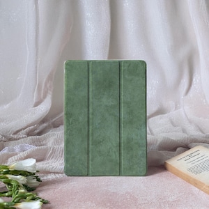 May include: A sage green tablet case with a textured finish. The case is rectangular with a tri-fold design. The background includes a book and white fabric.