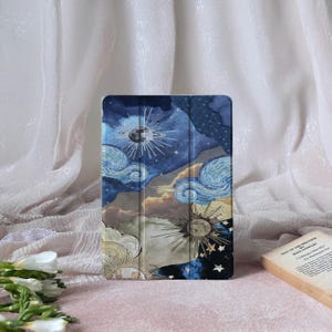 May include: A tablet case with a celestial design, featuring a night sky, sun, moon, and stars. The case has a blue, gold, and white colour scheme. The design is reminiscent of a painting. The case is rectangular and has a folding cover.