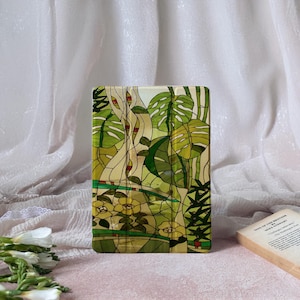 May include: A rectangular case with a stained glass design featuring green tropical leaves, yellow flowers, and tree trunks. The case has a cream-colored background and is surrounded by a white fabric.