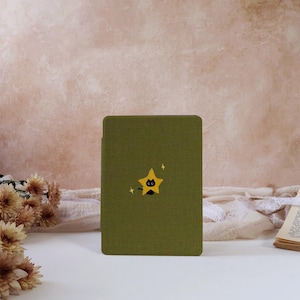 Op de afbeelding: Een olijfgroene e-reader hoes met een centraal geplaatste gele stergrafiek met daarin het silhouet van een kleine zwarte kat. De hoes heeft een textiele afwerking met textuur en is versierd met twee kleine gele vierpuntige sterren.