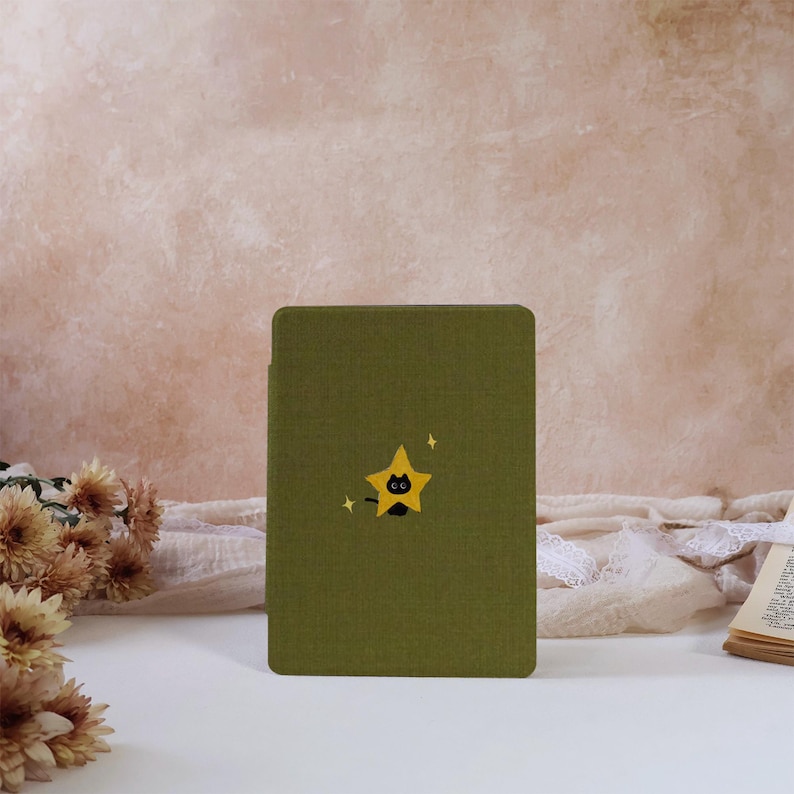Peut inclure: Une housse de liseuse rectangulaire vert olive orn&eacute;e d'un motif d'&eacute;toile jaune centr&eacute; contenant la silhouette d'un petit chat noir. Le design comprend deux minuscules &eacute;toiles jaunes &agrave; quatre branches de chaque c&ocirc;t&eacute; du motif central.