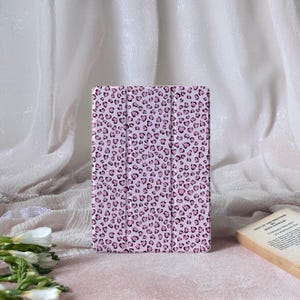 Puede incluir: Una funda rosa para tableta con un diseño de estampado de leopardo en forma de corazón. La funda está cerrada y tiene una cubierta magnética. El fondo es de un color suave y neutro.