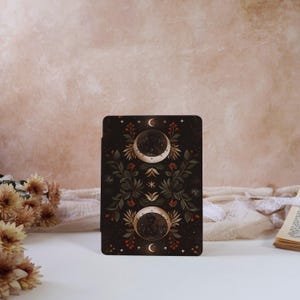 Peut inclure: Une carte rectangulaire foncée avec un motif céleste. Elle présente deux croissants de lune et une étoile centrale, ornée de motifs floraux et de baies vertes, rouges et dorées. Le fond est d'une teinte neutre et douce.