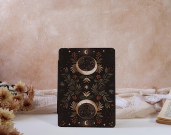 May include: A rectangular, dark-colored card featuring a celestial design with two crescent moons and a central star. The card is adorned with floral and berry motifs in shades of green, red, and gold. The background is a soft, neutral tone.