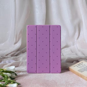 Może przedstawiać: Lawendowe etui na tablet z wzorem w kropki. Etui jest otwarte, odsłaniając wnętrze. Tło obejmuje białą tkaninę i książkę z tekstem.