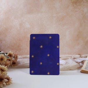 Puede incluir: Una tarjeta rectangular azul marino con patrones de estrellas doradas. La tarjeta está de pie sobre una superficie blanca, con flores secas y un libro en el fondo. El diseño evoca un tema celestial.