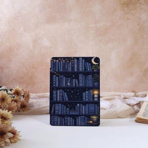 Puede incluir: Una funda rectangular para lector electrónico con una ilustración de estantería azul oscuro. Los estantes contienen libros, un faro, una planta en maceta, una taza de té y una luna creciente. El diseño incluye estrellas y luces centelleantes.