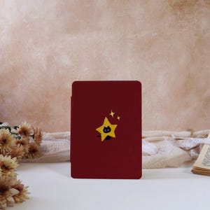 May include: A rectangular dark red e-reader case featuring a centered graphic of a small black cat peeking out from behind a bright yellow five-pointed star, accompanied by two small yellow star-shaped sparkles positioned above the main star.