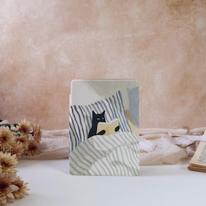 Peut inclure: Une coque de tablette décorative avec une illustration d'un chat noir lisant un livre au lit. Le design présente une couverture et un oreiller rayés dans des tons gris et bleus. Le chat tient un livre jaune.