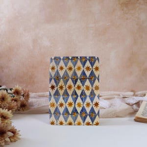 May include: A rectangular box with a blue and white diamond pattern, adorned with gold star accents. The box is standing upright on a white surface, with dried flowers and a book visible in the background. The box has a vintage aesthetic.
