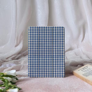 Può includere: Una custodia per tablet a quadretti blu e bianchi, su uno sfondo drappeggiato. Il motivo a quadretti è in stile vichy. Un libro intitolato "Sins of the Brother" è parzialmente visibile.