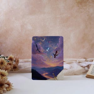 Puede incluir: Una funda rectangular para lector electrónico con una ilustración de fantasía de tres dragones volando sobre un paisaje montañoso al atardecer. La escena incluye una luna creciente, un cielo estrellado, nubes moradas y un río dorado. Una espada descansa sobre un libro.