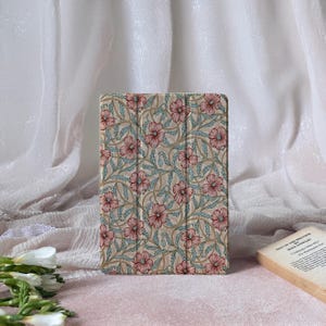 Puede incluir: Una funda para iPad con un estampado floral. La funda presenta un fondo beige con un diseño repetido de flores rosas, hojas azules y enredaderas verdes. La funda está cerrada y parece estar hecha de un material duradero.