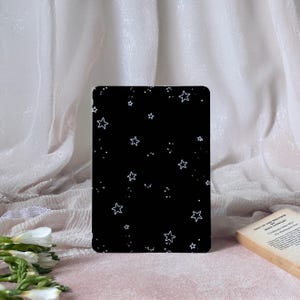 May include: A black rectangular case featuring a white star design. The case is displayed against a backdrop of soft, white, draped fabric. A book titled "Sins of the Brother" is partially visible.