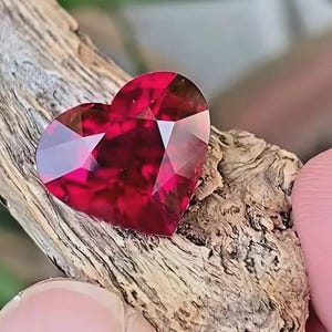 May include: A faceted, heart-shaped gemstone in a deep red hue. The gem is resting on a piece of weathered wood, showcasing its brilliant cut and reflective surfaces. The gemstone's facets catch the light, creating a sparkling effect.